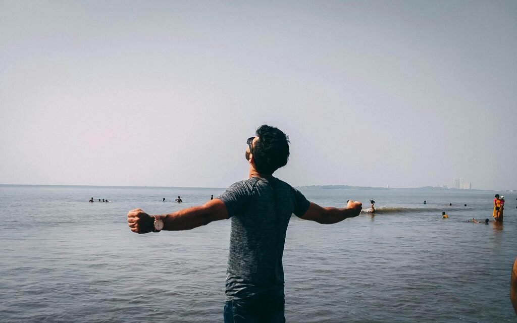 A man with open arms embracing freedom on a sunny beach day, surrounded by people enjoying the water.